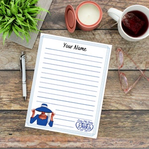 May include: A lined notepad with the text "Your Name" at the top and a blue border. A graphic of a woman in a blue hat and dress is in the bottom corner, along with the text "Zeta Phi Beta 1920". A pen, glasses, and a mug of tea are nearby.