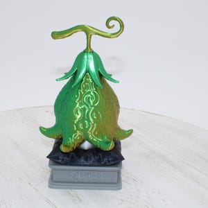 May include: A green and gold decorative fruit sculpture with a stem and leaf design. The sculpture is on a small, gray pedestal with text. The fruit is resting on a black pillow.