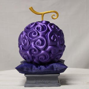 May include: A purple, spherical object with a swirling pattern, topped with a gold stem. It rests on a small, dark blue pillow and a gray pedestal. The design is reminiscent of a fantasy fruit.