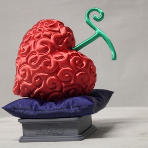 May include: A red heart-shaped fruit with a green stem, sitting on a blue pillow and a gray base with the word "ONE PIECE" printed on it.