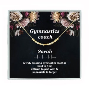May include: A black gift box with a floral design and gold trim. The box contains a gold necklace with the word "Sarah" spelled out in beads. The text on the box reads "Gymnastics coach... A truly amazing gymnastics coach is hard to find, difficult to part with & impossible to forget."