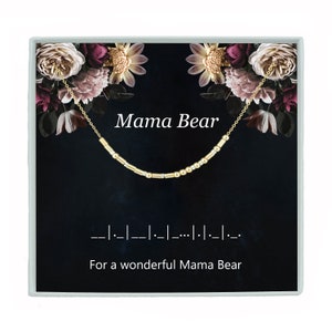 May include: A gold necklace with a floral design on a black background. The necklace has small gold beads and the words "Mama Bear" are printed on the box. The text "For a wonderful Mama Bear" is also printed on the box.