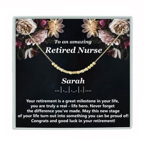 May include: A gold necklace with a nameplate that reads "Sarah" and a message that says "To an amazing Retired Nurse". The necklace is displayed in a black box with a floral design.