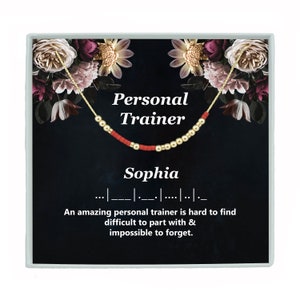 May include: A gold-colored necklace with red and gold beads, presented in a gift box. The box features the words "Personal Trainer" and "Sophia", along with a Morse code pattern and a motivational quote about personal trainers.