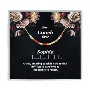May include: A black and gold gift box with a floral design. The box contains a gold necklace with red beads and the text "Best Coach Ever Sophia". The text below the necklace reads "A truly amazing coach is hard to find difficult to part with & impossible to forget."