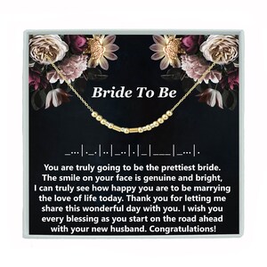 May include: A gold necklace with a beaded charm that says "Bride To Be" on a black background with floral accents. The text reads: "You are truly going to be the prettiest bride. The smile on your face is genuine and bright, I can truly see how happy you are to be marrying the love of life today. Thank you for letting me share this wonderful day with you. I wish you every blessing as you start on the road ahead with your new husband. Congratulations!"