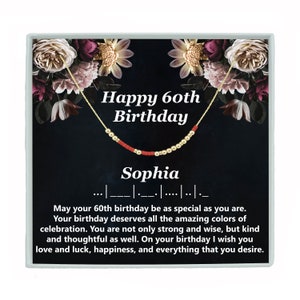 May include: A black and gold gift box with a floral design. The box contains a gold necklace with red beads and the text "Happy 60th Birthday Sophia".