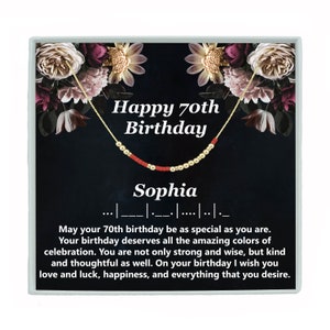May include: A black and gold gift box with a red and gold beaded necklace. The box has a floral design and the text "Happy 70th Birthday Sophia" on the front. The text "May your 70th birthday be as special as you are. Your birthday deserves all the amazing colours of celebration. You are not only strong and wise, but kind and thoughtful as well. On your birthday I wish you love and luck, happiness, and everything that you desire." is printed on the bottom of the box.