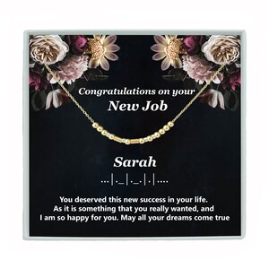 May include: A gold necklace with a beaded chain and the word "Sarah" spelled out in beads. The necklace is in a black box with a floral design and the text "Congratulations on your New Job".