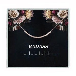 May include: A gold necklace with a beaded chain and the word "BADASS" written in white text on a black background with a floral design.