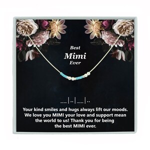 May include: A silver necklace with turquoise and white beads, presented in a black box with a floral design. The box features the text "Best Mimi Ever" and a message about kind smiles and hugs.