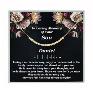 May include: A gold necklace with a beaded chain and a nameplate with the name "Daniel" on it. The necklace is in a black box with a floral design and the words "In Loving Memory of Your Son".