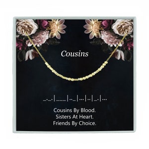 May include: A gold necklace with small beads and a charm that says "Cousins". The necklace is in a black box with a floral design and the text "Cousins By Blood. Sisters At Heart. Friends By Choice."