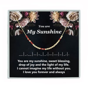 May include: A gold necklace with red beads and the words "You are My Sunshine" on a black background with floral accents.