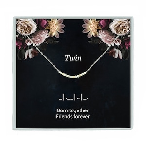 May include: A silver necklace with a delicate chain and a charm that reads "Twin" in silver lettering. The charm is made up of small silver and gold beads. The necklace is displayed in a black box with a floral design and the text "Born together Friends forever".
