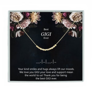 May include: A gold necklace with a bar chain and small gold beads spelling out "Best Gigi Ever" on a black background with floral accents. The necklace is in a white gift box.