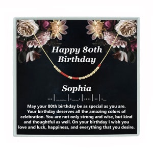 May include: A black and gold gift box with a floral design. The box contains a red and gold beaded necklace with the text "Happy 80th Birthday Sophia" on the front.