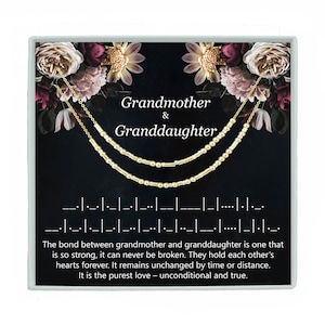 May include: A gold necklace set for a grandmother and granddaughter, presented in a gift box. The necklaces feature a Morse code design. The box has floral accents and the words "Grandmother & Granddaughter".