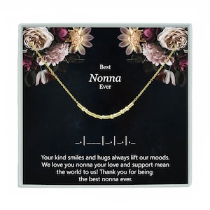 May include: A gold necklace with a message that reads "Best Nonna Ever" in a black box with a floral design. The necklace is made of small gold beads and is displayed on a black background.