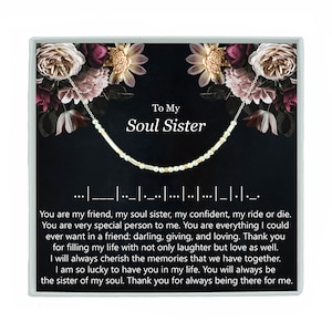 May include: A silver necklace with a message in Morse code that reads "To My Soul Sister". The necklace is displayed in a box with a floral design and a black background.