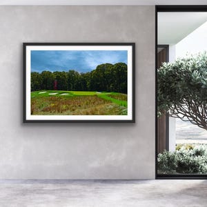 以下が含まれることがあります： ゴルフコースの額入り写真。画像には、緑のフェアウェイ、バンカー、遠くの赤い構造物が写っています。空は青とグレーの色合いで曇っています。写真は灰色の壁に表示されています。