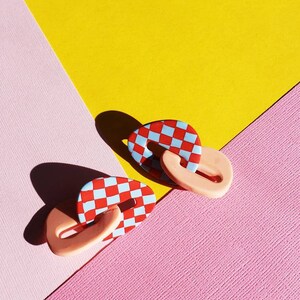 May include: A pair of red and blue checkered oval shaped earrings with a pink accent. The earrings are on a pink and yellow background.