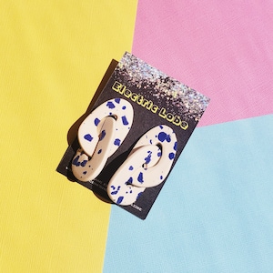 May include: A pair of white ceramic earrings with blue splatter paint. The earrings are shaped like a loop and are on a black card with the text "Electric Lobe".