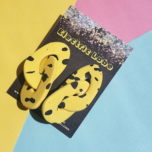 May include: A pair of yellow and black patterned earrings with a chain link design. The earrings are on a black card with the text "Electric Lobe".
