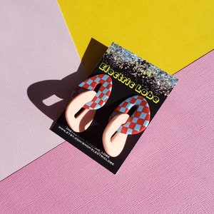 May include: A pair of red and blue checkered earrings with a pink loop on each side. The earrings are on a black card with the text "Electric Lobe" and a website address.