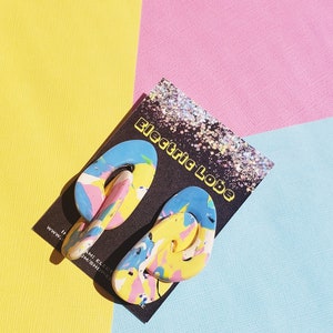 May include: A pair of colorful abstract swirl earrings with blue, yellow, pink, and white paint. The earrings are on a black card with the text "Electric Lobe" in white.
