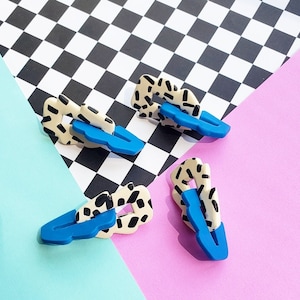 May include: Four decorative hair clips. Each clip has a blue base and a cream-colored top with black speckles. The clips are arranged on a background of black and white checkerboard, light blue, and pink.