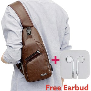 May include: A brown leather crossbody bag with a zipped pocket and a silver buckle. The bag is being worn by a person. The image also shows a pair of white earbuds in a white package with the text "Free Earbud".