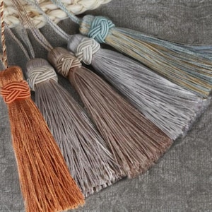 Op de afbeelding: Close-up van vijf decoratieve kwasten in tinten oranje, bruin, grijs en blauw. Elke kwast heeft een geknoopt bovenstuk en een lange, vloeiende franje. De kwasten zijn diagonaal geplaatst tegen een grijze achtergrond.
