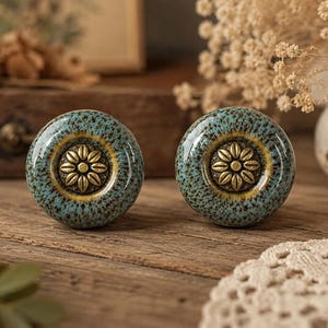 Puede incluir: Dos pomos redondos de cerámica para armarios con un esmalte moteado en tonos azul verdoso y marrón. Cada pomo tiene una incrustación central de metal color latón con un diseño floral detallado de doce pétalos en patrón radial.