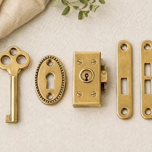 May include: A collection of antique brass door hardware, including a skeleton key, an oval escutcheon, a rectangular lock, and two elongated plates with rectangular cutouts. The items are displayed on a light surface.