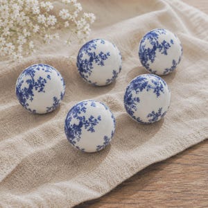 Puede incluir: Cinco esferas de cerámica blanca con un patrón floral azul están dispuestas sobre una superficie de lino beige. Las esferas están decoradas con un delicado diseño botánico azul. El arreglo está junto a un pequeño ramo de flores blancas.