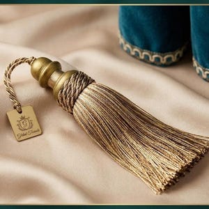 May include: A decorative gold and brown tassel with a tag that reads "Gilded Threads". The tassel has a braided top and a long, flowing fringe. The tag is rectangular with a crown and the letter "G".