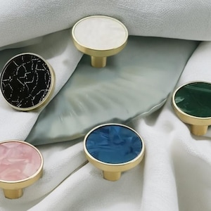 Peut inclure: Une collection de boutons de meubles décoratifs avec des bases en laiton et des dessus marbrés colorés. Les boutons présentent une variété de couleurs, dont le blanc, le noir, le rose, le bleu et le vert. Les boutons sont ronds et ont un design simple et élégant.