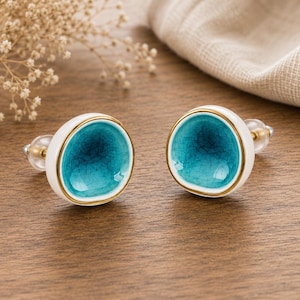 Peut inclure: Deux boutons de meuble en céramique. Chaque bouton présente une base blanche, un bord doré et un centre turquoise craquelé. Les boutons ont un dos transparent à visser. Les boutons sont sur une surface en bois.