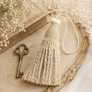 May include: Cream-colored tassel with a woven top and a long, braided fringe. A decorative key and a vintage book are also visible. The tassel is hanging on a decorative tray, with a sprig of white flowers.