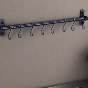 May include: A black metal kitchen utensil rack with eight curved hooks. The rack is mounted on a neutral-coloured wall. The design is simple and functional, suitable for hanging pots, pans, or other kitchen tools. The rack is approximately 61 cm long.