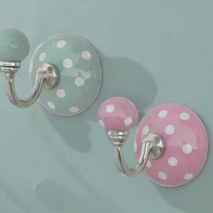 Könnte beinhalten: Zwei dekorative Wandhaken. Einer ist mintgrün mit weißen Punkten, der andere rosa mit weißen Punkten. Beide haben einen silberfarbenen Haken und einen runden Knopf.