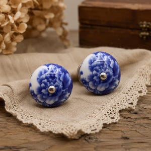 May include: Two round ceramic cabinet knobs featuring a white base with a blue floral pattern. Each knob has a smooth, glossy finish and a small, centred silver-toned metal screw head on the front, providing a functional hardware element.