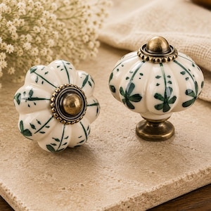 Puede incluir: Dos tiradores decorativos para armarios. Uno tiene forma de flor, el otro de calabaza. Ambos son blancos con diseños de hojas verdes y detalles en latón. Los tiradores están sobre una superficie beige, con flores blancas en el fondo.
