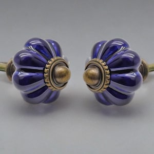 Może przedstawiać: Dwa dekoracyjne gałki do szafek. Każda gałka ma błyszczący, głęboki niebieski, żłobiony ceramiczny wzór z mosiężną metalową podstawą i śrubą. Gałki są przeznaczone do łatwej instalacji.