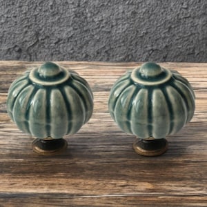 May include: Two teal ceramic cabinet knobs with a ribbed, pumpkin-like design. Each knob has a small, round top and a bronze-colored base. The knobs are displayed on a wooden surface.