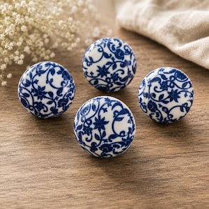 Puede incluir: Cuatro pomos redondos de cerámica con base blanca y diseño floral azul. Los pomos están dispuestos sobre una superficie de madera, mostrando su diseño decorativo. La combinación de colores azul y blanco añade un toque clásico. Cada pomo mide unos 4 cm de diámetro.
