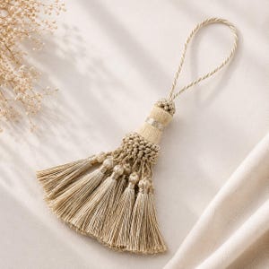 May include: A beige tassel with a loop for hanging. The tassel features a decorative beaded section, a silver band, and multiple strands of fringe. The design is symmetrical and the color is neutral.