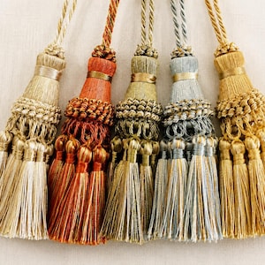 May include: Five decorative tassels in beige, rust, olive green, grey-blue, and gold. Each tassel has a braided top, a decorative band, and a long fringe. The tassels are arranged side by side.