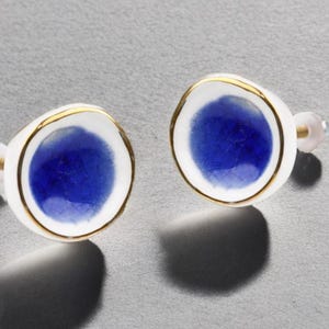 Puede incluir: Un par de pendientes de botón con un diseño de cerámica blanca y borde dorado. Cada pendiente tiene un centro azul vibrante. Los pendientes están sobre un fondo gris, mostrando su elegante diseño.